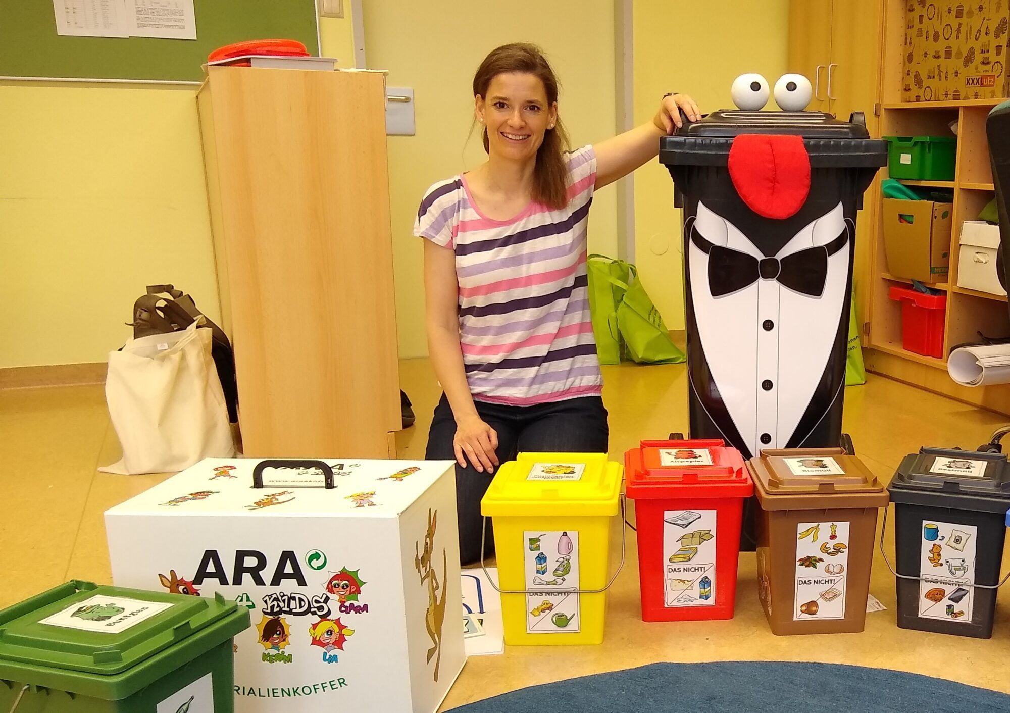 Zwei Kinder im Volksschulalter stehen neben einer bloden Frau mit geflochtenen Zöpfen und einer Schleife im Haar. Davor ein Siegespodest mit Abfällen darauf und im Hintergrund eine Tafel und ein Regal mit blauen Ordnern. 
