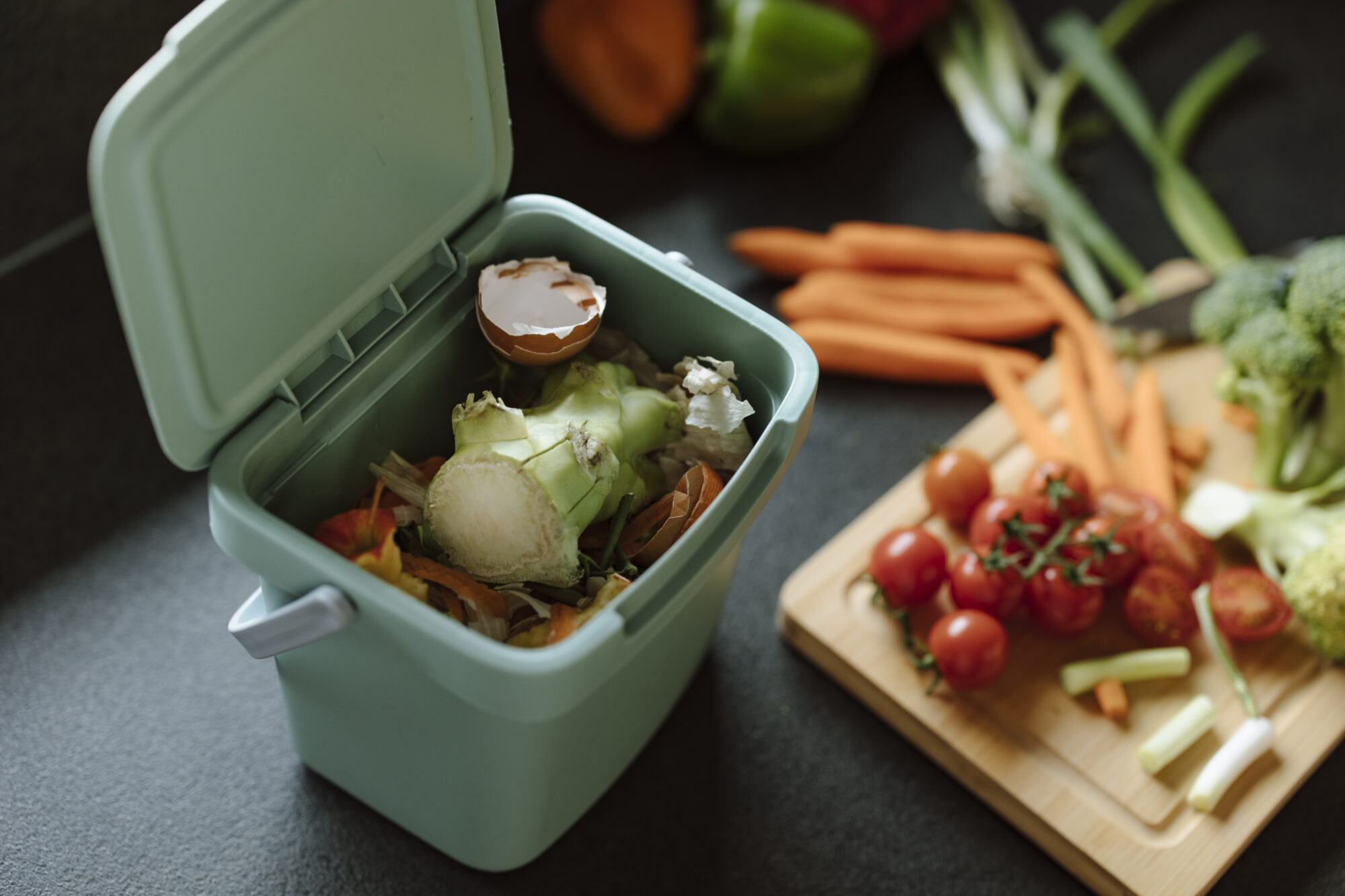 Auf der rechten Seite ist ein kleiner grüner Abfallbehälter mit Gemüseabfällen zu sehen. Links davon ein Scheidebrett aus Holt mit Tomaten, Karotten, Jungzwiebel und Brokkoli.